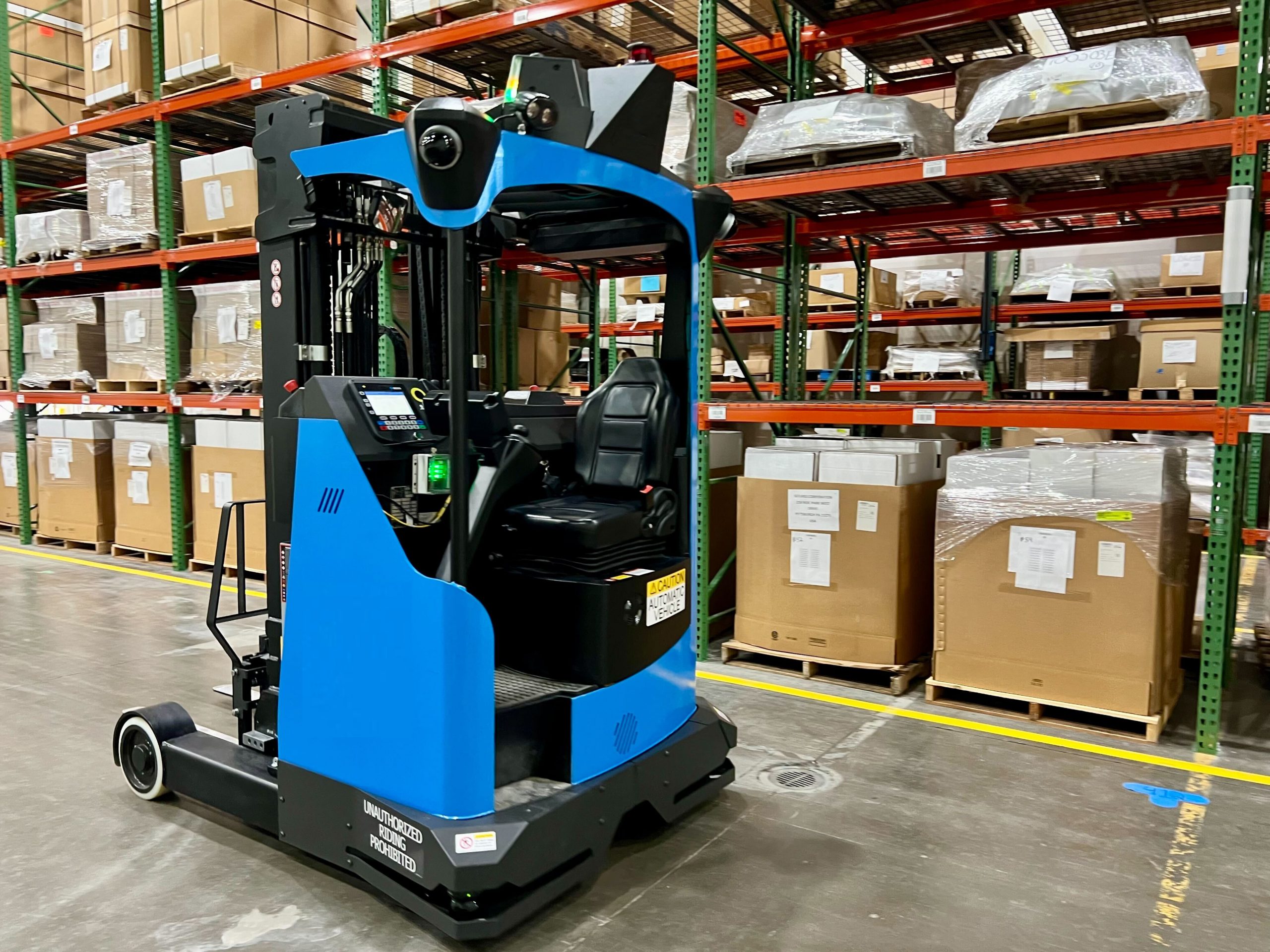 Blue and black forklift in a warehouse, positioned in front of multi-level shelving filled with large boxes. Yellow floor markings indicate pathways and storage zones.