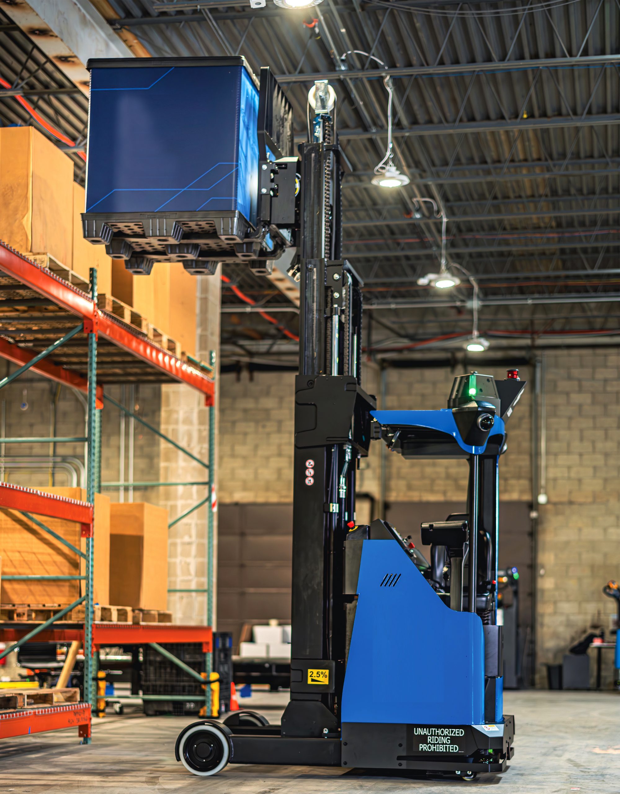 Automated blue and black forklift lifting a large blue container in a warehouse with high ceilings, shelving, and visible safety signage.