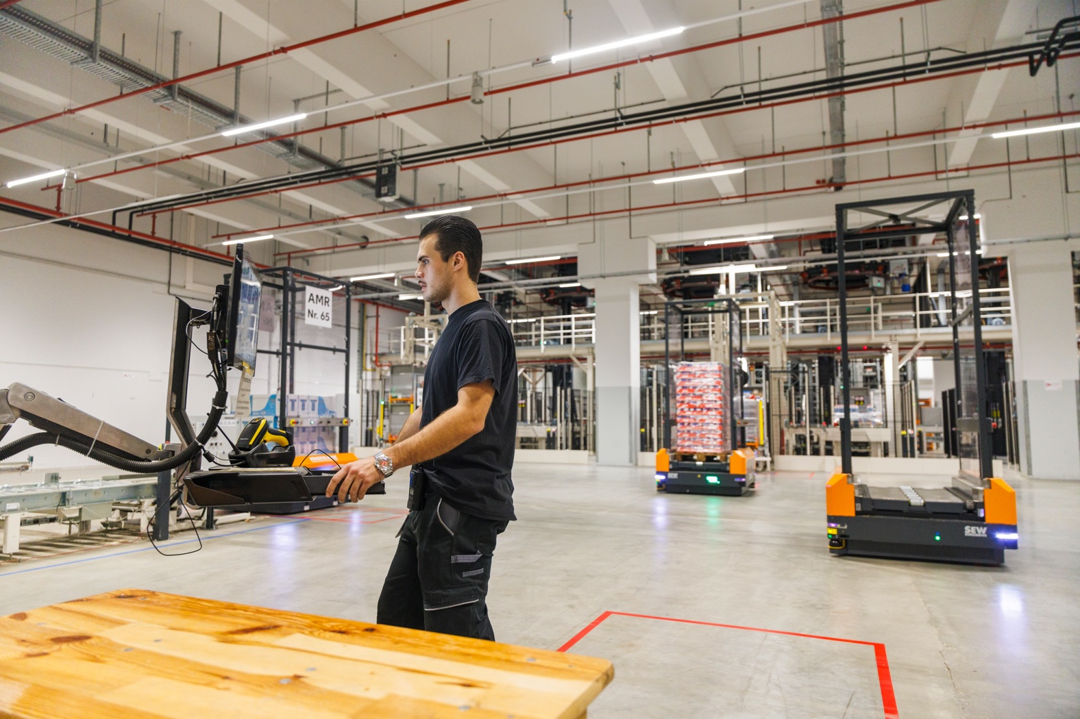 Person operating machinery in a warehouse, holding a control device while monitoring equipment. Automated guided vehicles (AGVs) transport goods in the background. The warehouse features high ceilings with exposed pipes and lighting.